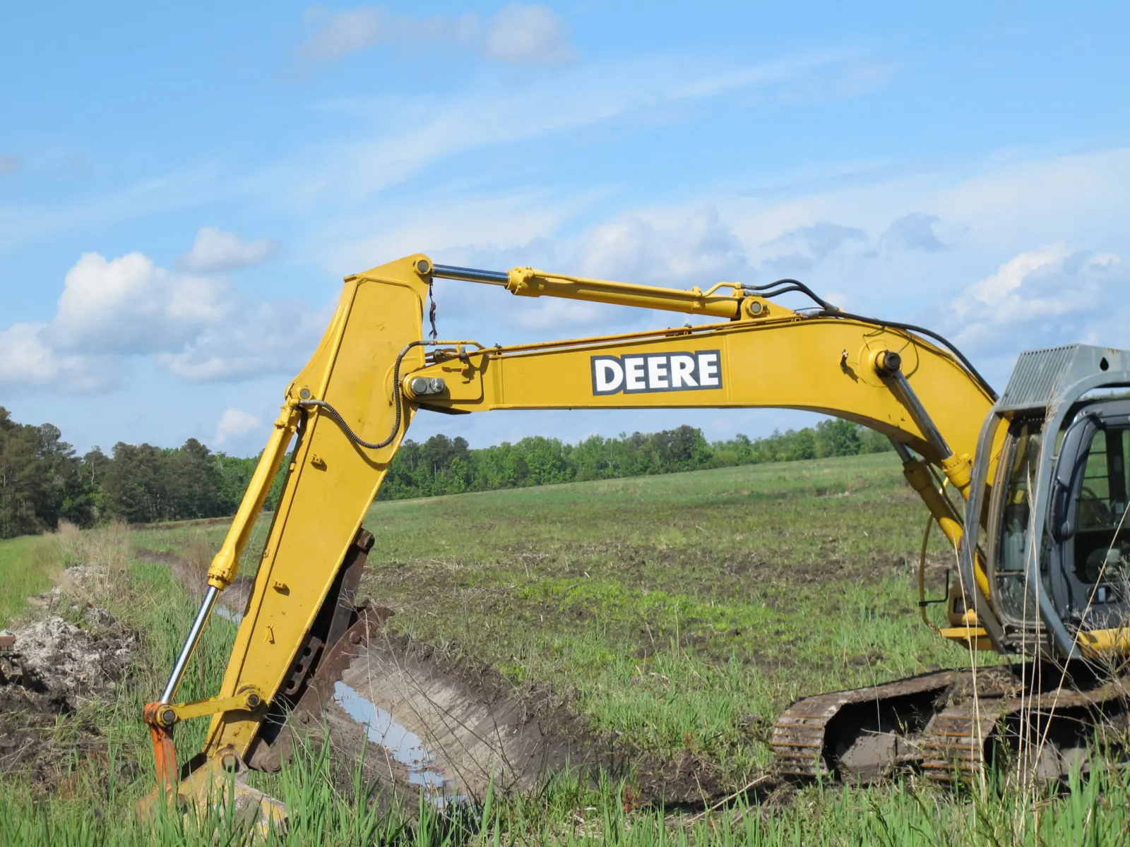Excavator boom cutting a drainage ditch through a green farm field near pines