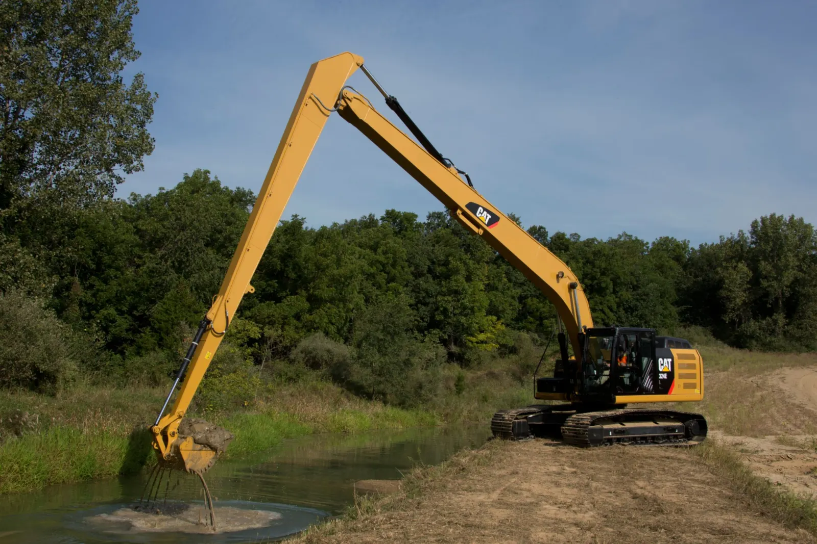 Long reach excavator clearing muck and vegetation from a pond bank beside wooded shoreline