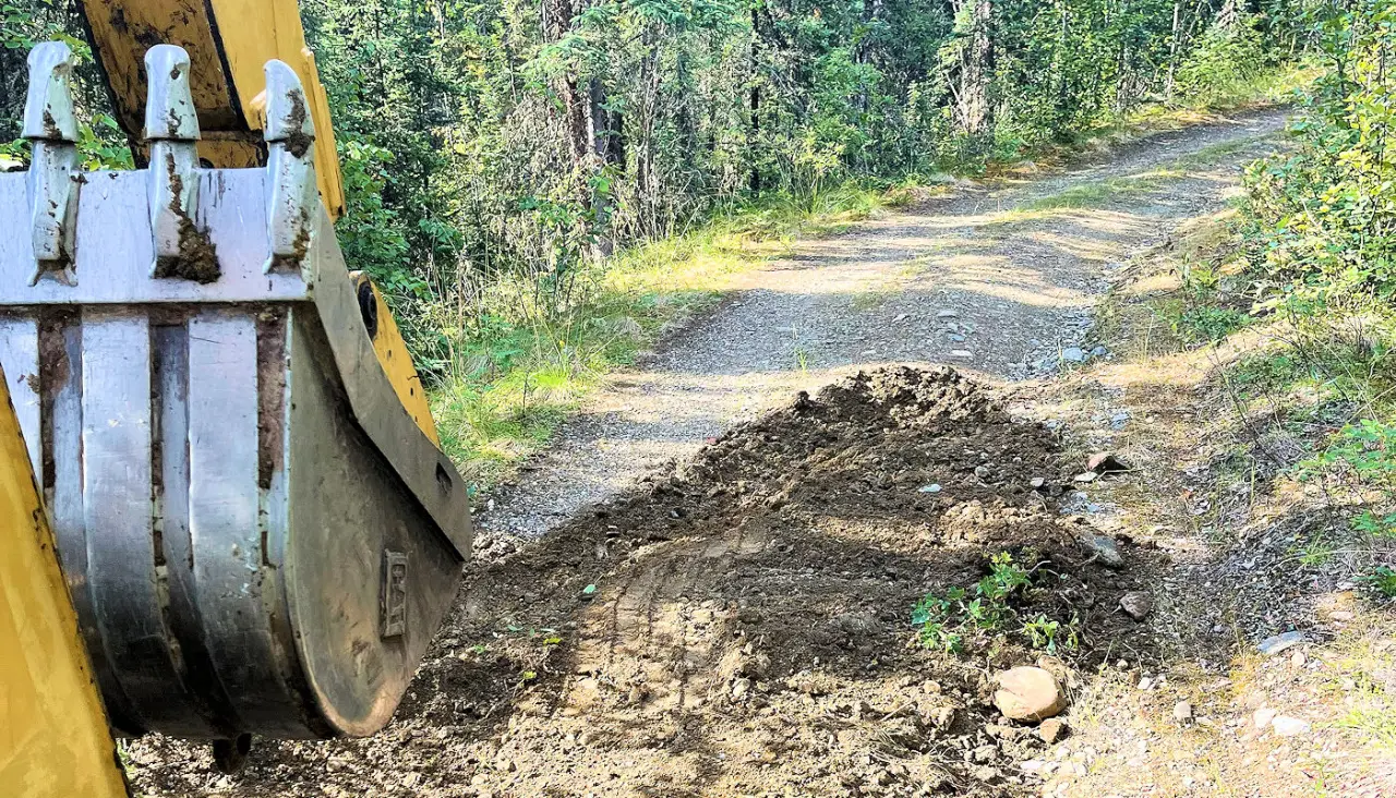 Excavator bucket spreading base material while building a gravel farm road through woods