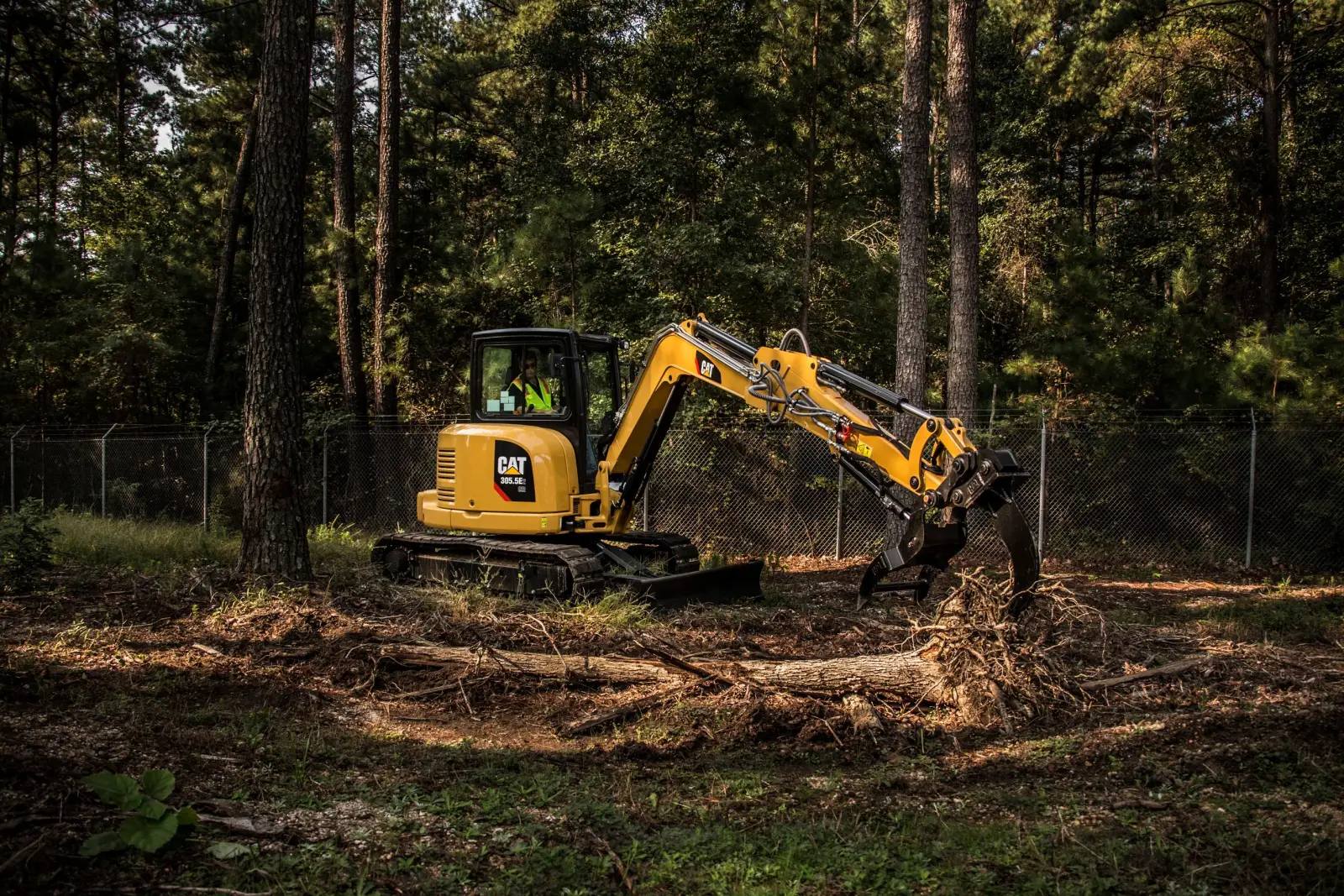 Compact excavator with grapple thumb pulling logs while clearing a wooded lot among pines